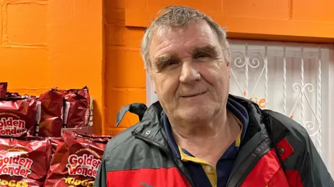 A man smiles as he stands in front of piles of red bags of Golden Wonder crisps inside the community shop. A brick wall behind him is painted orange, while a window is covered with white bars in a trellis design.