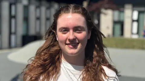 Robyn is smiling at the camera. She has long curly reddish-brown hair resting over her shoulders. She is wearing a white T-shirt with a delicate gold necklace visible over the top. She is standing in front of the college building.