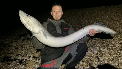 Tre Hart A man in black and grey fishing clothing holding a long grey eel.
