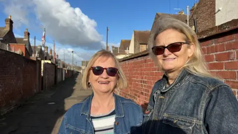 Andrew Turner/BBC Jenny Butcher, standing left, and Linda Osborne. They're both wearing denim jackets and sunglasses, and they both have blonde hair. They stand in an alley – which stretches into the distance behind them – between two rows of terraced houses. 