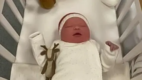 A tiny infant in a crib with white sheets and bars. She is hoding her hands up in the air and has a white baby sleeper on and white knitted cap with a red strip. there is a toy reindeer under her right arm