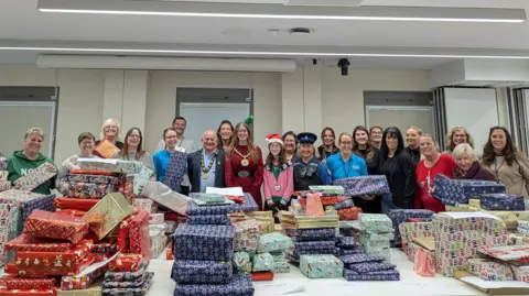 Epping Forest District Council A row of proud-looking people of different ages and uniforms obscured by a big, multi-coloured pile of presents which stretches the whole way across the row of people