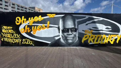 Steve Jenkins Mural of Carl Cox on baordwalk by stadium. Cox is a black man with shaved head. he has been painted wearing sunglasses in black and white with yellow letters either side saying Oh yes oh yes, and The Prodigy 