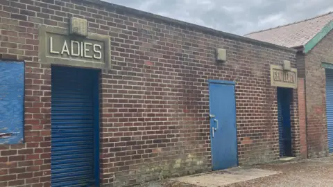 Picture of the former ladies and mens changing rooms for Otley Lido. The words MENS and WOMENS are etched in stone above two blue doors on a bricked wall.