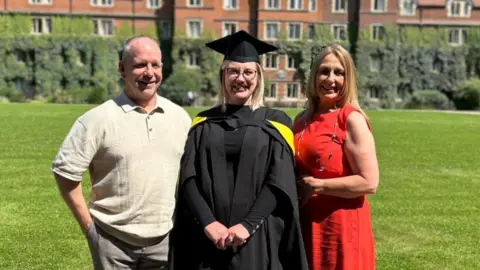 Handout Three people standing in front of a lawn of an old building. Charlotte's father on the left with little hair on his head and wearing a polo shirt. Charlotte in the middle wearing all black graduation robes including a mortarboard on her head. Joanna Donohoe on the right wearing a red dress. All three are looking at the camera on a sunny day, smiling.