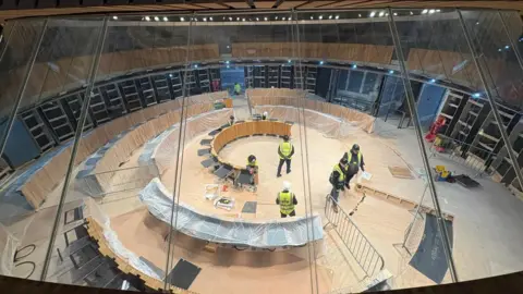 BBC The image shows construction workers at work in the Senedd debating chamber. The politicians' desks are covered in polythene.