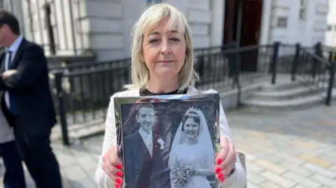 Collette Rooney has light blond hair down to her shoulders and is holding a black and white wedding photograph of her dad and mum while standing outside court