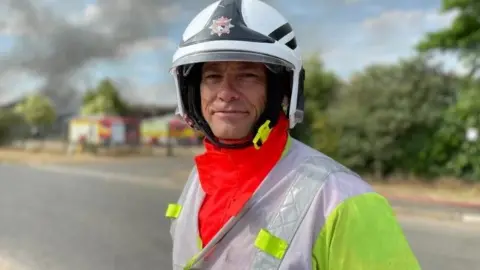 Shaun Whitmore/BBC A male firefighter poses for a photograph in his helmet and uniform with fire engines in the background