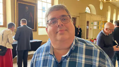 A man with short grey hair and glasses, wearing a check shirt containing blues of various shades.