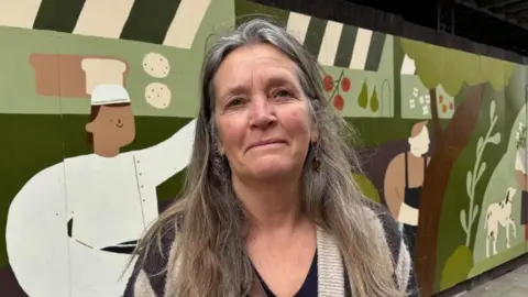 Charlie Stubbs / BBC There is a woman with grey long hair, with a brown and grey cardigan and a black T-shirt. She has earrings in both ears with a silver loop. She has a colourful artwork of people cooking and someone collecting a bouquet of flowers with a dog