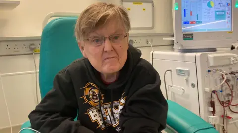 The image shows a man in a hospital setting, wearing a black Boston Bruins hoodie, while he receives kidney dialysis.