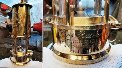 Supplied Two images of the same restored mining lamp sitting on a paper towel in a shed. The lamp is very shiny. The left image shows the full lamp. The left image is close up of the base. It is engraved with the words “Les Bauser. Retired miner. Bolsover Colliery. 19.10.83”