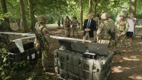 Dan Jarvis, a man in a suit, speaks with soldiers dressed in camouflage, outdoors, surrounded by trees.