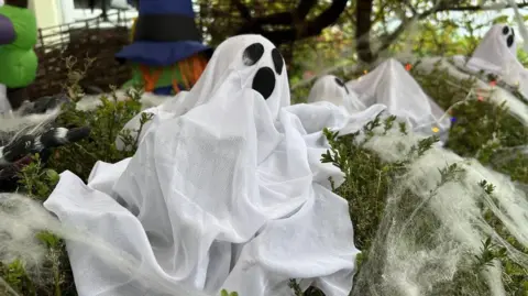 A spooky white ghost with large black eyes and mouth sat on a green hedge covered with white spider webs. 