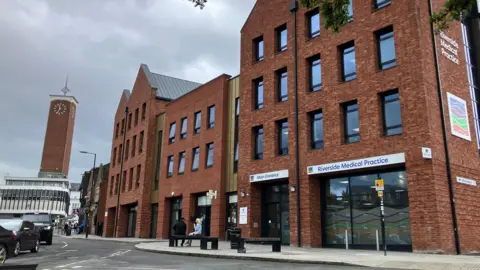 A four-storey red-brick building with both flat and pitched roofs. A glass entrance can be seen on the right hand side of the building with a sign reading "Riverside Medical Practice". A tall bricked clock tower and road can be seen in the distance.