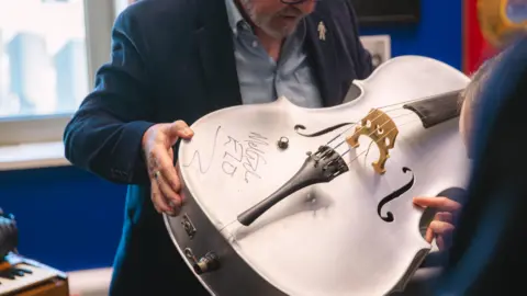 Birmingham City University A man holds a large silver cello, which has been autographed.