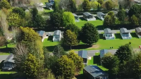 An aerial view of Allerthorpe holiday park featuring multiple cabins arranged in neat rows. Each unit is surrounded by lush green lawns, with a mix of evergreen and deciduous trees scattered throughout the area. Paved pathways connect the houses, and some units have vehicles parked beside them. The grass appears freshly trimmed.