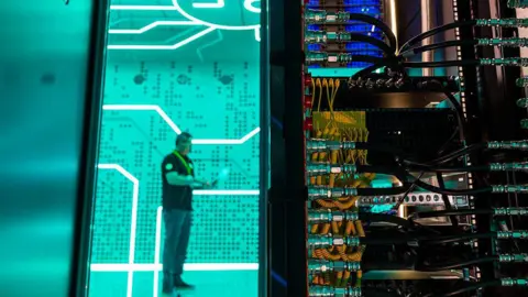A man standing against a bright green background in a data centre - in the foreground is the back of a large computer with lots of yellow and black wires.