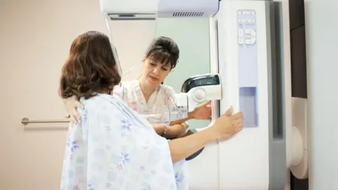 Getty Images A woman in a faintly patterned hospital gown holds on to a piece of hospital equipment as she prepares for a mammogram. She is facing away from the camera, towards a healthcare worker who is helping adjust her position.