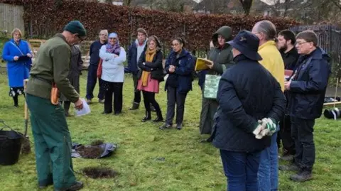 North Northamptonshire Council A wide shot photo of 12 people stood on a cold winter day. They are watching a man demonstrate how to plant a tree.