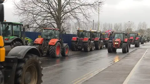 Tractors, mostly red, travel in both directions down a road on a grey and damp day. One of them is carrying a Union flag. Bare trees and street lamps can be seen in the background.