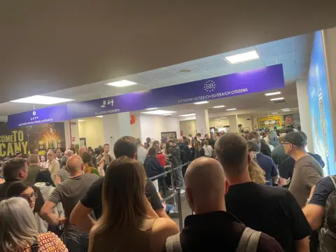 John McKenzie Heavy queues at passport control in Pisa Airport. The signs for EU and non-EU travellers can be seen.