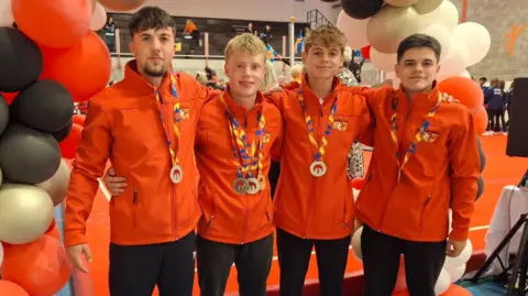 Helen Duggan Four young male gymnasts wear red coats and dark trousers and stand under a balloon arch with their arms around each other. They have medals on ribbons around their necks. One with blonde hair in the centre has four medals.