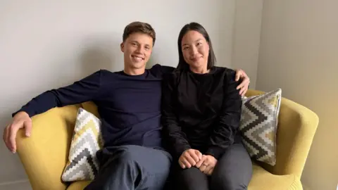 A woman and a man sit next to each other on a yellow sofa. The woman has shoulder-length black hair and wears a black jumper and dark trousers. The man has his arm around the woman's shoulders. He has short brown hair and is wearing a navy jumper. They are both smiling and look relaxed.