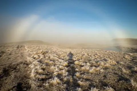 SAIS Southern Cairngorms A sweeping moor with a covering of frost. The fogbow appears to arch above the silhouette of the person photographing it.