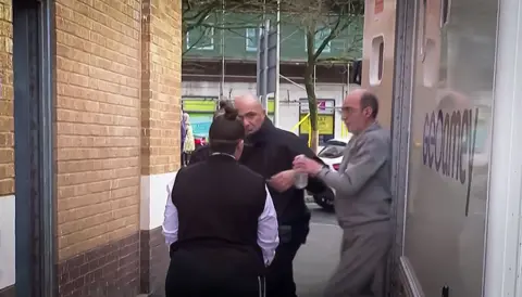 BBC Anthony Felton is being led down the stairs of a police custody van. Two officers, a bald man and a woman with her hair in tight top bun guide Felton towards the door of a brick building, Swansea magistrates court. He has handcuffs on his hands which are stretched out in front of him with a plastic water bottle in one hand. he has male pattern baldness with no hair on top and short cropped hair at the sides. He has a light beard and is wearing a grey track suit top and bottoms