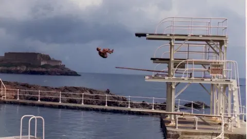 Andre Quevâtre A man diving from an outdoor diving board into a pool in Guernsey. The man is holding his shins and in the middle of a flip. There are cliffs in the background to the left and the sea behind the pool.