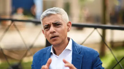 Reuters Sadiq Khan is seen sitting wearing a light suit. He is gesturing with his hands while speaking and an urban green space is visible behind him.