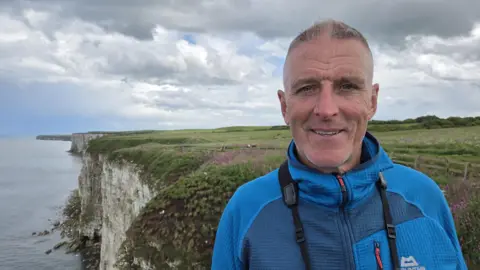 A man with short cropped grey hair, wearing a bright blue outdoors jacket, stands on the top of a tall, chalk cliff with fields and the sea in the background. 