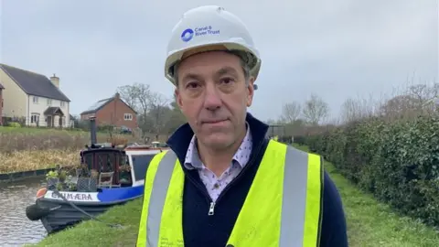 A man with a white hard hat and a yellow hi-vis jacket. he is standing next to a canal and a narrowboat