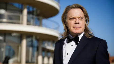 Getty Images Suzy Eddie Izzard stands outdoors in formal black tie evening wear. The modernist architecture of the De La Warr Pavilion with its large glass windows and white balconies is blurred in the background.