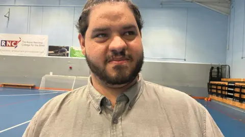 A man in a grey shirt standing inside a sports hall, with the Royal National College for the Blind sign visible behind them.