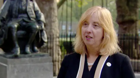 Dr Carol Homden, a woman with shoulder-length blonde hair, wearing a dark jacket with a string of pearls, standing outside Coram's headquarters near a statue of Thomas Coram, who started the Foundling Hospital