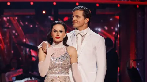 BBC Amber Davies and Nikita Kuzmin in a photo from the semi final on the strictly dance floor. She is wearing a white sequin dress and has her long brown hair tied back. He is wearing a white suit. they are holding hands and he has his hand on her shoulder 
