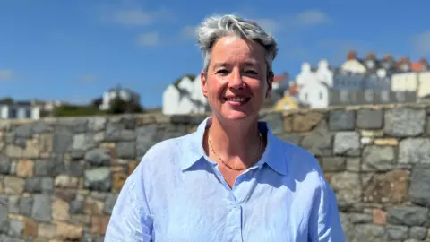 Woman in her 40s wearing a blue shirt with houses and a stone wall in the background