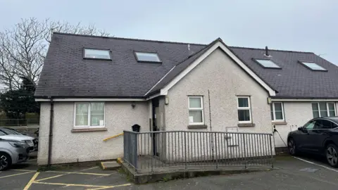 Cambria Surgery, viewed from its car park. It is a single storey building with a pebbledashed outside and noticeable skylights in the slate roof. The gable end of a porch faces the camera with access ramp leading across the front of it.
