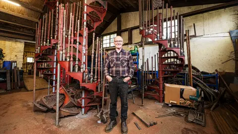 Mark Lewis Paul Dennis wears a checked shirt, black jeans and glasses in his workshop, a stone outbuilding. He is smiling directly at the camera. Behind him are two sets of metal spiral staircases. Individual metal steps are piled beneath one of the spirals. 