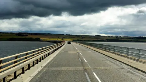 Looking down the carriageway of the Cromarty Bridge. There is one car in the distance. The firth's water are grey, reflecting dark clouds above.