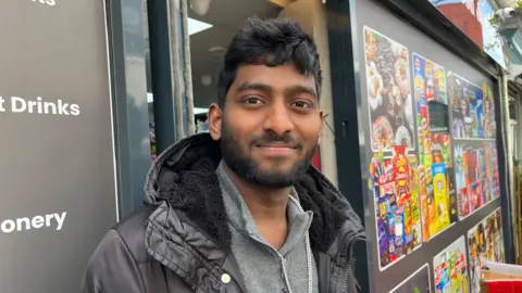 Dineshkumar Ravisekar is looking into the camera and smiling. He is wearing a grey fleece with a black hooded coat over the top. He is standing in the doorway of a shop. Over his left shoulder there is a collage of brightly coloured food packaging.