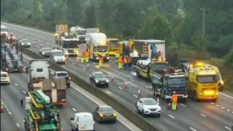 National Roads A CCTV image of the section of the M25. You can see a single lane line of traffic going through the only open road. Next to it are several large trucks and people in high-vis.