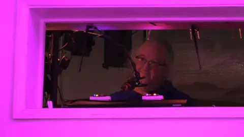 Looking in at Charles in his sound booth, framed by the window and pink lighting.