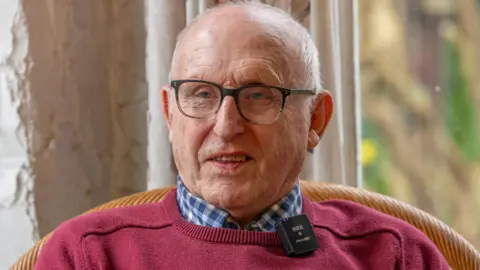 Dressed in a salmon jumper and a blue and white checked shirt, woth short grey haor and glasses, Hirzel Dorey smiles at something just off-camera. He has microphone on his collar as he is being interviewed.