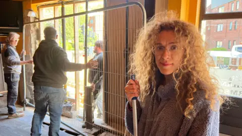 Emily Rogers looks at the camera holding a broom. She has long curly hair and glasses and wears a purple fleece. Men install barriers to cover the damaged wall behind her. 
