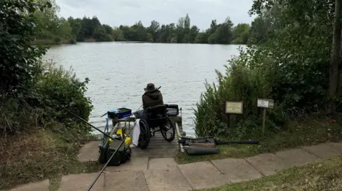 Tony Prior fishing, pictured from the back. He is beside the lake and is on a base near a paved area. 