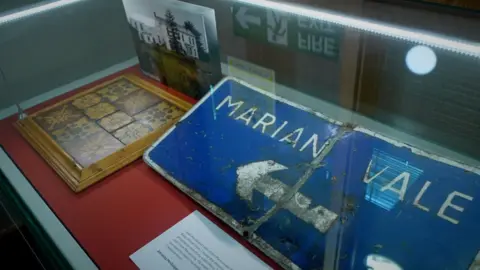 Exhibits on display in The Sunflower Project in the Linenhall Library, a Marian Vale sign in a cabinet alongside tiles and a photograph of the former home.