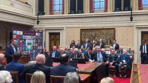 People sitting in Stormont buildings. A man is speaking at the front of the room.